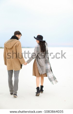 Happy woman and man walking on snow and holding hands, wearing coat, grey scarf and hat. Concept of seasonal inspiration and couple photo session.