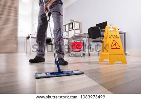 Low Section Of Male Janitor Cleaning Floor With Caution Wet Floor Sign In Office Royalty-Free Stock Photo #1083973499