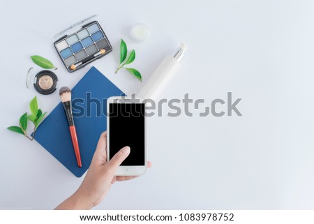 woman hand holding smart phone and blue book under brush with eyeshadow makeup and green leaves near serum bottle skin care on white background with copy space