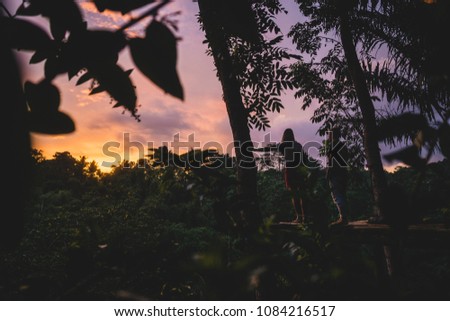 Two female travellers sitting on a bamboo platform between trees looking out over the jungle while watching the sunset in Tetebatu, Lombok, Indonesia