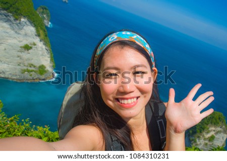 young beautiful and happy Asian Korean tourist woman taking selfie picture smiling carrying backpack after hiking excursion on top of tropical sea cliff landscape in holidays travel destination