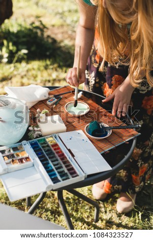 Brush in the hands of the artist, watercolor painting, creativity. Watercolor painting with brush at the easel. An artist paints a landscape on an easel in a flowering garden