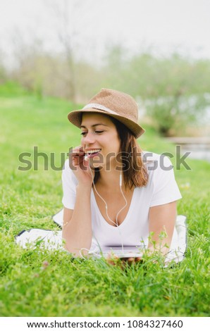 Young happy woman lying in the grass and enjoying music from her phone in the nature. Smiling. Wearing hat.