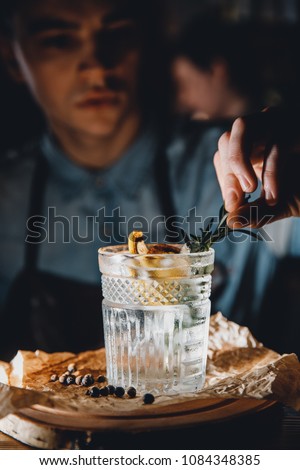Barman prepares cocktail with orange and herbs in transparent glass on bar with alcohol. Uses tongs for decoration. Dark background. Royalty-Free Stock Photo #1084348385