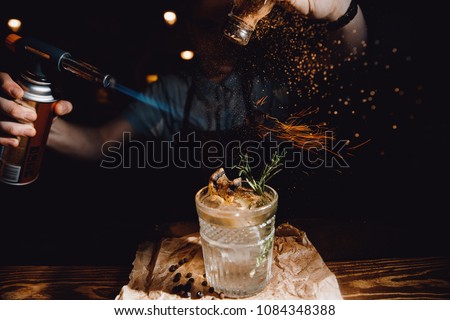 Barman prepares cocktail with orange and herbs in transparent glass on bar with alcohol. Uses burner with sparks. Dark background. Royalty-Free Stock Photo #1084348388