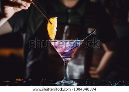 Barman prepares cocktail with orange and martini purple color on bar with alcohol. Uses tongs for decoration. Dark background. Royalty-Free Stock Photo #1084348394