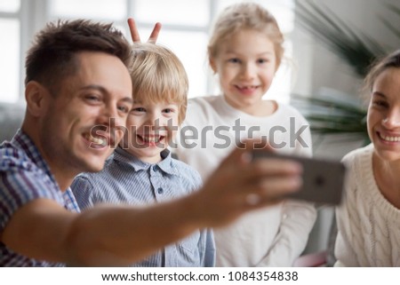 Kid sister making little cute brother bunny ears while father taking happy family selfie, funny children boy and girl having fun posing for self-portrait with smiling mom and dad bonding together
