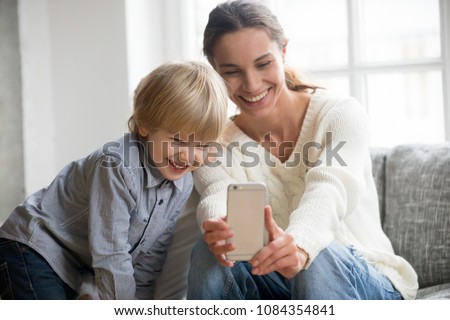 Happy mother laughing taking selfie with little son on smartphone at home, smiling single mom and cute adopted boy playing making photo posing for self portrait, mommy and kid watching video on cell