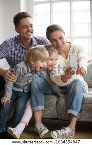 Happy mother taking family selfie with husband and kids looking at smartphone, playful adopted children siblings having fun posing for funny photo on phone with smiling mom and dad together, vertical