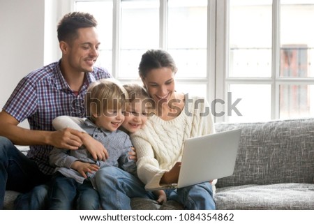 Young family of four bonding using laptop sitting on sofa at home, happy parents with adopted cute kids son and daughter having fun with computer watching video or doing internet shopping together