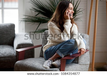 Happy cheerful young woman talking on the phone at home, smiling teen girl making answering call by cellphone sitting on chair, beautiful lady having pleasant funny conversation speaking by mobile Royalty-Free Stock Photo #1084354877