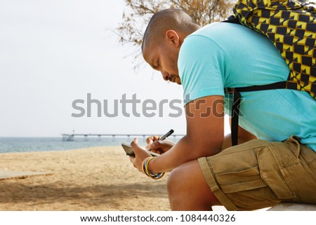 Pensive African-American hipster man wearing backpack and making notes in copybook outside of the city. Student preparing for lesson at college on the ocean beach