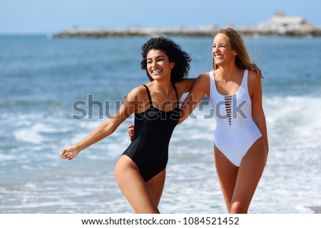 Two young women with beautiful bodies in swimwear on a tropical beach. Funny caucasian and arabic females wearing black and white swimsuits walking along the shore.