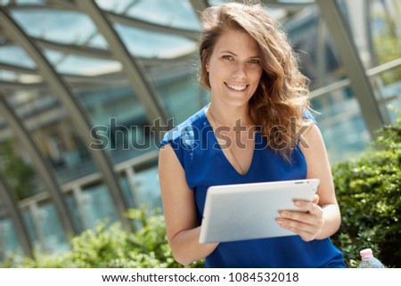Young woman holding tablet, sitting outdoors, smiling happy.