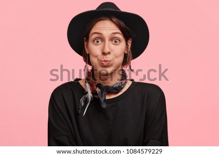 Beautiful stunned female blows cheeks in surprisment, dressed in black clothing, as talk with friend, isolated over pink background. Adorable cowgirl shocked to find out about winning racing