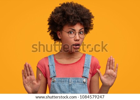 Discontent African American female makes stop sign with palms, disagrees to do something, demonstrates her refusal, poses against yellow background. Dark skinned young woman shows no gesture Royalty-Free Stock Photo #1084579247