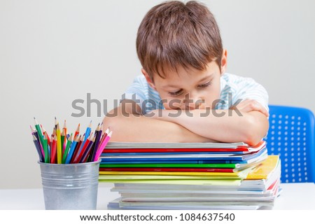 Upset schoolboy sitting at desk with pile of school books and notebooks