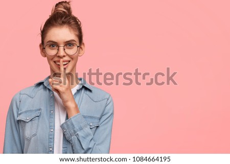Pleased attractive young woman with happy smile, shows hush gesture, asks to keep confidential information in secret, poses against pink studio background with copy space for your advertisement