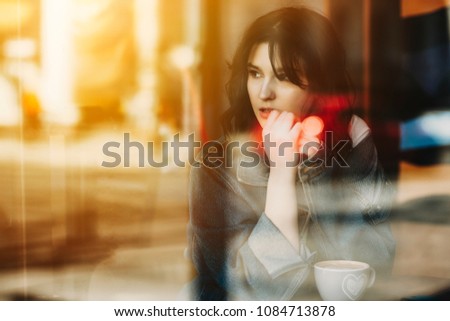 Image through the window of a attractive short haired woman sitting in a coffee shop and looking away touching her lips.