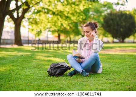 Beautiful girl sitting on grass and reading book at the park in summer. Student girl is studying in a park at sunset.