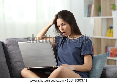Shocked teen reading news online in a laptop sitting on a couch in the living room at home