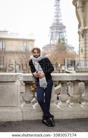 Afro american handsome young man standing near concrete railing with Eiffel Tower background in Paris. Boy wearing grey scarf and black sweater. Concept of fashionable look and trip to France.