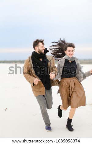 Happy young man with beardand woman running on snow and wearing fashionable clothes, winter background. Concept of seasonal photo session and romantic relationship.