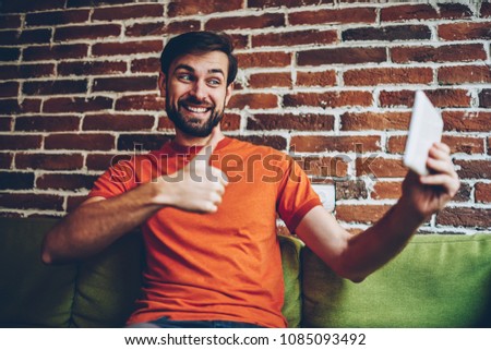 Joyful bearded male blogger in orange t-shirt making selfie photo on front camera of digital tablet.Positive hipster guy with thumb up shooting video on modern touch pad connected to 4G internet