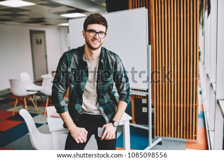 Half length portrait of handsome successful male designer wearing glasses for provide eyes protection enjoying break time at work and looking at camera while sitting on table in modern office