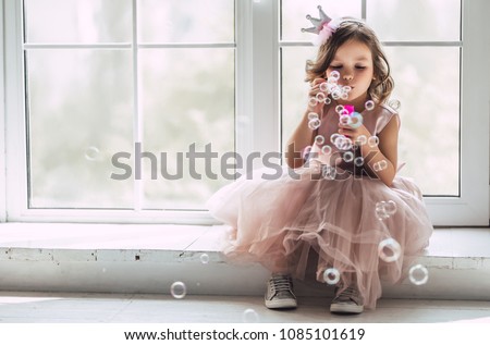 Little cute girl in beautiful dress is sitting near the window at home and blowing soap bubbles. Royalty-Free Stock Photo #1085101619