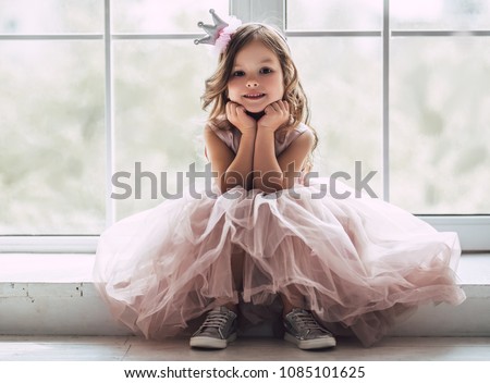 Little cute girl in beautiful dress is sitting near the window at home. Royalty-Free Stock Photo #1085101625