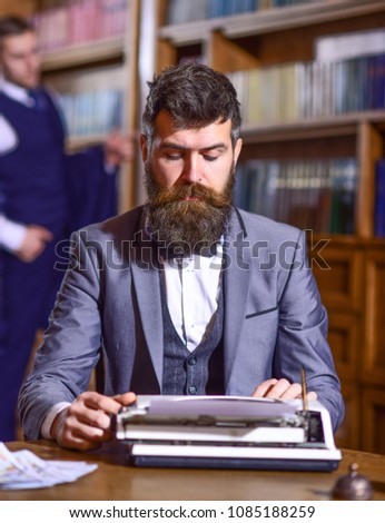 Man with beard and busy face sit in library and work with typewriter, close up. Writers routine concept. Author types novel or poem. Writer working on new book with bookshelves on background.