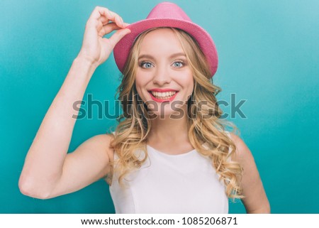 A picture of happy woman wears pink hat. She is holding the edge of ger pink hatwith one hand. Girl is cheerful. Isolated on blue background.