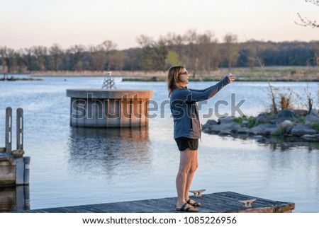 woman standing on pier taking photo of sunset with smartphone