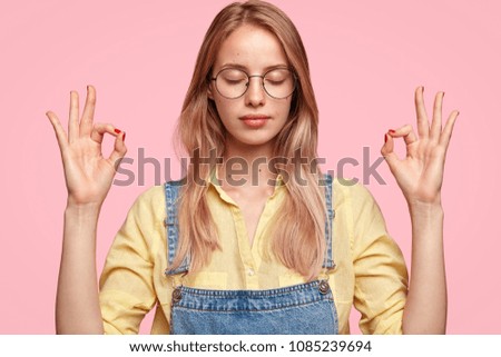 Calm beautiful young female tries to relax after hard working day at office, holds hands in mudra gesture, keeps eyes shut, wears stylish clothing. Student feels peaceful and relaxed, practices yoga
