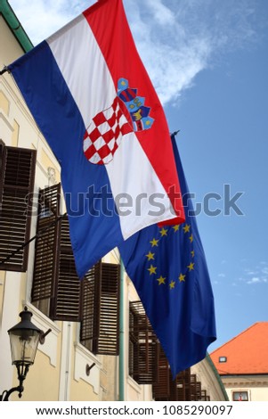 Flags of Croatia and European Union hang out together on wall of building with open shutters, in background red roof of another building, blue sky