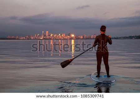 Adventurous girl on a paddle board is paddeling in the ocean with Downtown City in the background during a vibrant sunset. Taken near Spanish Banks, Vancouver, British Columbia, Canada.
