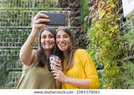 two beautiful women taking a selfie with mobile phone. One is holding a cup of coffee. They are laughing. Outdoors lifestyle and friendship concept