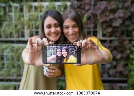 two beautiful women taking a selfie with mobile phone. One is holding a cup of coffee. They are laughing. Outdoors lifestyle and friendship concept