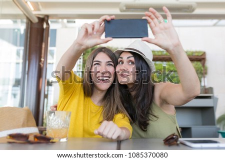 two beautiful women having breakfast in a restaurant and taking a selfie with mobile phone. They are laughing. Indoors lifestyle and friendship concept