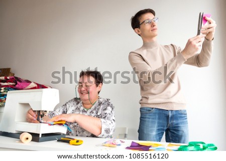 patchwork, creativity, art concept. in the light workshop there is a comand of dressmakers, old smiling woman is sitting at the desk and stitiching with sewing machine and a young man is helping her