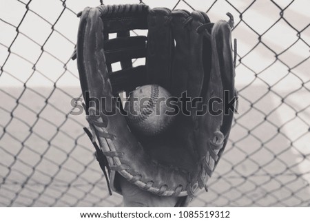 American sport of baseball shows closeup of ball in black and white with glove.  Dugout fence in background