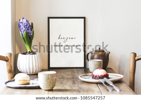 Stylish interior design of kitchen space with small table with mock up frame, plant, cups of tea and tasty dessert.