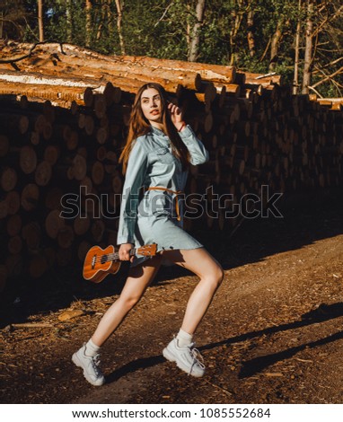 Young attractive girl is walking in the woods at sunset in a long blue shirt with ukulele in hands
