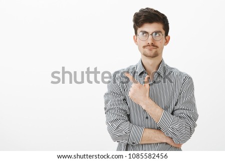Portrait of indifferent unsurprised nerdy guy with moustache, pointing and looking at upper left corner with tight smile, being displeased with no care about topic, standing over gray background