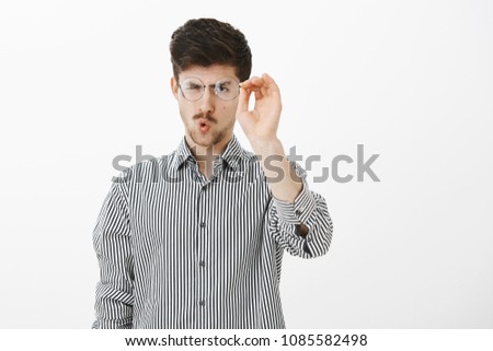 Portrait of ordinary adult european male student, taking off glasses and cleaning glass of eyewear, staring at goggles focused, folding lips to blow, standing over gray background