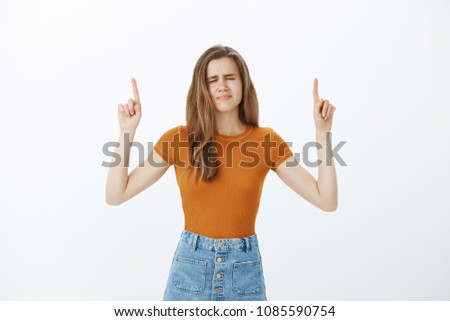 Intense nervous attractive woman in stylish outfit, raising index fingers and pointing up with closed eyes and anxious expression, scared of spider on ceiling, waiting till in gone over gray wall