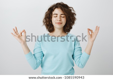Keep calm and carry on. Portrait of satisfied relaxed young caucasian woman with pleased smile, closing eyes and holding spread hands with zen gesture while meditating, mastering yoga over gray wall