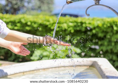 Closeup photo of young girl washing hands in city fountain with water splashing on them with a lot of drops, outdoors at sunny day, green hedge and lake on background