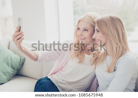 Stylish trendy modern cheerful mother and daughter having smart cell phone in hand shooting self portrait on front camera sitting on sofa in living room enjoying free time